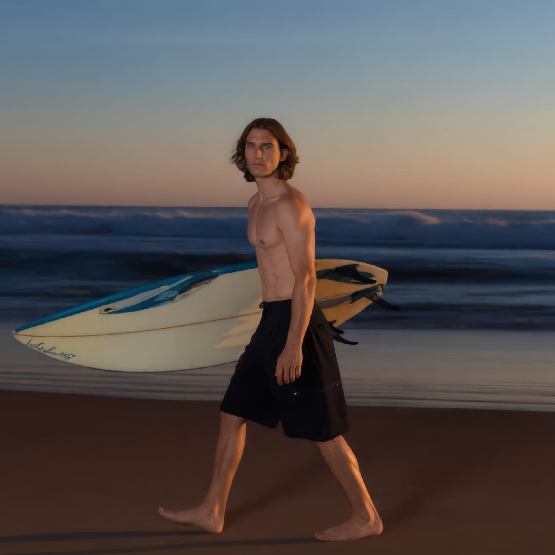 A surfer carrying a board along the shoreline at sunset, wearing the boardshort.